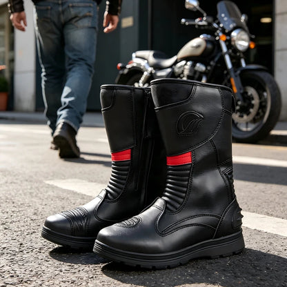 A pair of black leather motorcycle boots with red accents, displayed on an asphalt street with a blurred motorcycle and a person in jeans in the background.